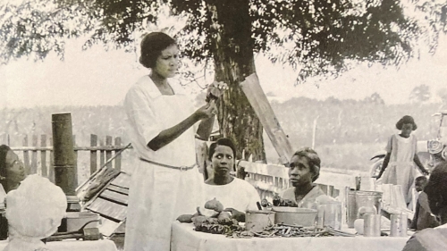 Black women sit at a table preparing food. One woman stands in the center; everyone is watching her. 