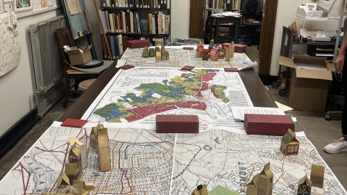 Image of a table covered by a large printed map. Small houses are constructed out of cardboard and other materials. 