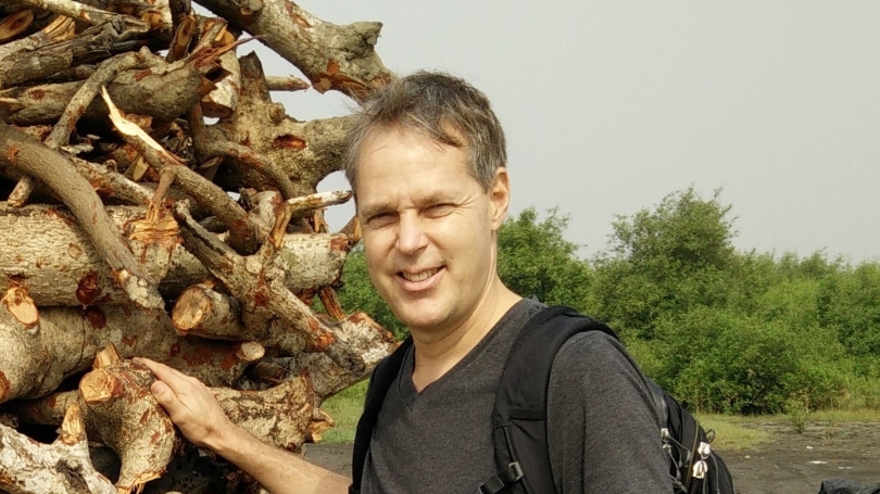 Alex smiles at camera in front of pile of logs