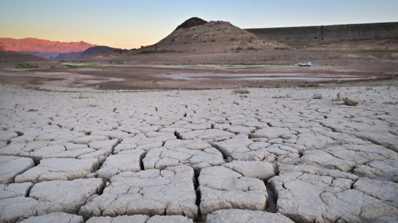 Image of dry, cracked lake bed of Lake Mead in the foreground, with a car driving by and hills in the background. 