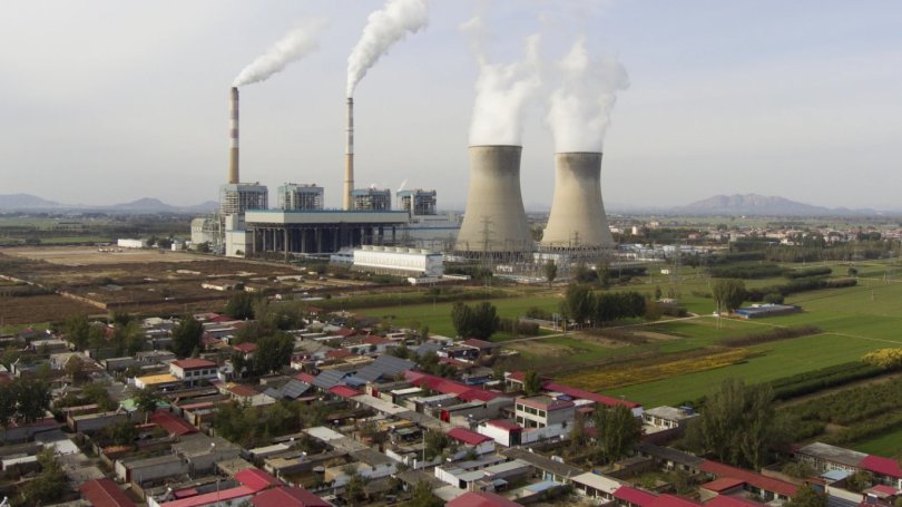 View of large coal-burning power plant surrounded by red-roofed houses, spewing smoke into a gray sky