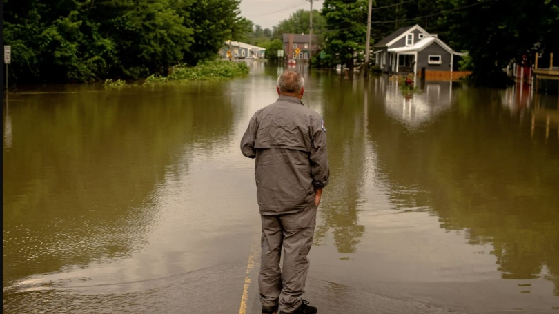 A person wearing a jumpsuit stands and looks out over the flooded road and houses, their back to the camera. 
