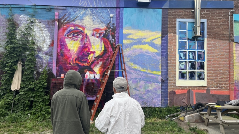 Two people stand facing the mural, a multi-colored image of a person's face and mountains.