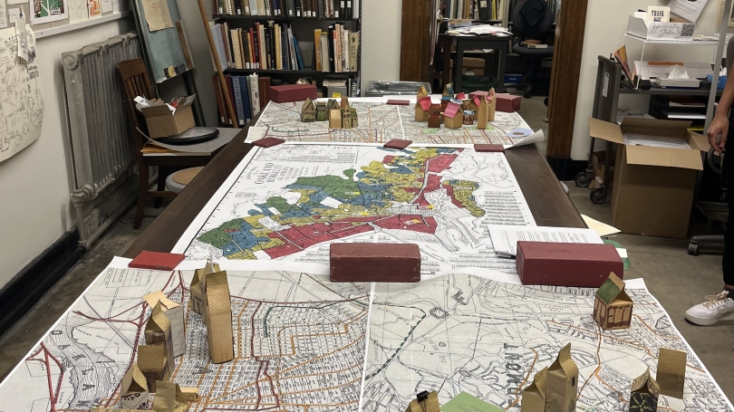 Image of a table covered by a large printed map. Small houses are constructed out of cardboard and other materials. 