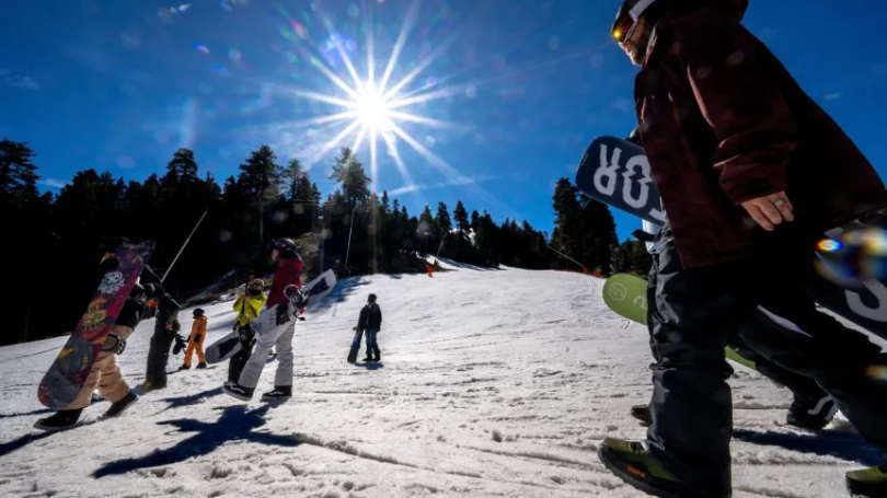 Person carries snowboard on ski mountain while sun shines bright overhead. 