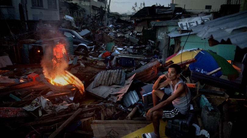 Man sits on top of destroyed house while another makes a fire. Street is full of splintered wood and roofing materials.