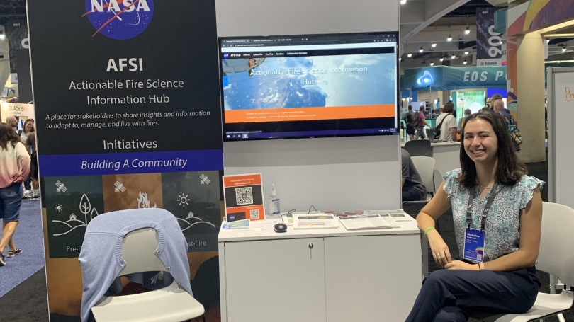 Madeline Duhnoski sits in front of a booth at a conference. The poster at the booth reads 'NASA AFSI: Actionable Fire Science Information Hub. A place for stakeholders to share insights and information to adapt to, manage, and live with fires.'